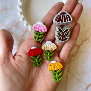 May include: Five plastic flower-shaped cookie cutters, including three different flower designs and two leaf designs. The cutters are in shades of red, yellow, white, and green.