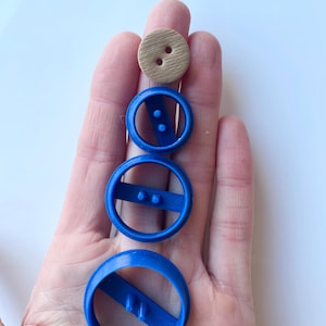 Könnte beinhalten: Eine Sammlung von Bastelartikeln: drei blaue Kunststoff-Kreisschneider und ein Holzknopf. Die Schneider haben einen Steg mit zwei erhabenen Punkten. Der Knopf ist hellbraun.
