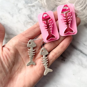 May include: Silver fish bone earrings with a textured finish, shown with two pink fish bone-shaped cookie cutters. The earrings hang from silver hooks. The background is a marble surface.