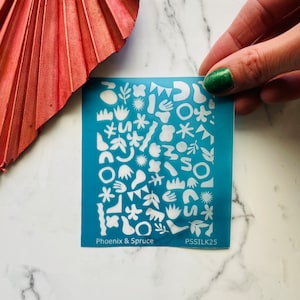 Peut inclure: Une feuille de pochoir bleu turquoise avec des formes découpées blanches de différents motifs abstraits. La feuille de pochoir porte l'inscription "Phoenix & Spruce" et "PSSILK25".