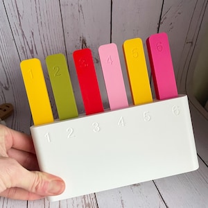 May include: A white rectangular organiser base with six slots, each labelled with a number from one to six. Six flat, rectangular markers in yellow, green, red, light pink, gold, and magenta are inserted into the slots, each marked with a corresponding number.
