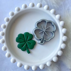 May include: A green four-leaf clover cookie cutter and a green four-leaf clover cookie on a white plate with a beaded edge.