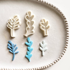 May include: Set of six white and blue leaf-shaped cookie cutters. The cutters are arranged on a white oval plate with a decorative edge.