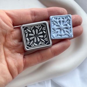 Puede incluir: Dos moldes cuadrados de silicona para manualidades. El molde más grande es plateado con un diseño floral detallado. El molde más pequeño es azul claro con el mismo diseño floral.