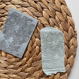 May include: Two gray polymer clay stamps with floral patterns are displayed on a woven, light brown surface. One stamp is rectangular, and the other is a curved shape. The stamps feature detailed designs of flowers, leaves, and other botanical elements.