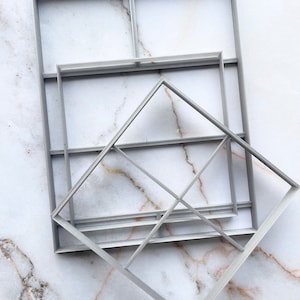 May include: A set of four gray plastic square cookie cutters, stacked on top of each other. The largest cutter is a square, the next is a rectangle, the next is a smaller square, and the smallest is a diamond shape.