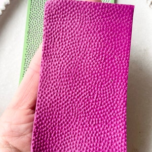 May include: A sheet of pink textured polymer clay with a raised dot pattern. The clay is being held in a hand.