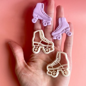 May include: Four white plastic roller skate cookie cutters on a pink background. Two of the cutters are purple.