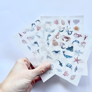 May include: Sheets of watercolor ocean-themed stickers featuring various marine life, including dolphins, whales, seahorses, and starfish. The stickers are in shades of blue, red, and beige, with a white background.