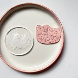 May include: A clear acrylic stamp with a floral design of pink flowers and leaves. The stamp is on a white plate with a pink rim.