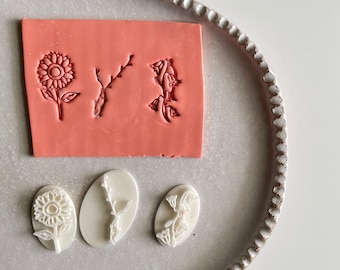 May include: A set of three white polymer clay stamps featuring floral designs. The stamps are on a white ceramic plate with a scalloped edge. The designs include a sunflower, a branch with leaves, and a cluster of flowers.