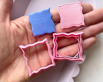 Può includere: Quattro stampini per biscotti, due rosa e due blu, a forma di quadrato con bordi arrotondati. Gli stampini sono su una mano.
