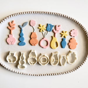 Puede incluir: Un conjunto de 15 cortadores de galletas en varias formas, incluyendo jarrones, flores y figuras abstractas. Los cortadores están hechos de plástico blanco y están dispuestos en un plato de cerámica blanco.