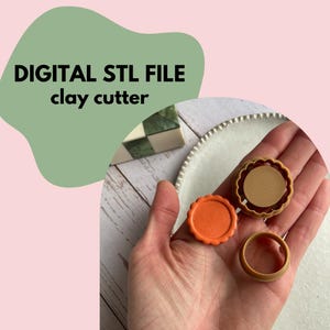 Op de afbeelding: De afbeelding toont drie kleisnijders in een hand, met de tekst "DIGITAL STL FILE clay cutter" in het zwart. Eén snijder is oranje en de andere twee zijn bruin. De snijders zijn cirkelvormig met decoratieve randen. De achtergrond is lichtroze en groen.