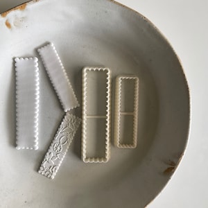 May include: Four white plastic cookie cutters on a white ceramic plate. The cutters are in the shape of rectangles with scalloped edges. Two of the cutters are larger than the other two.