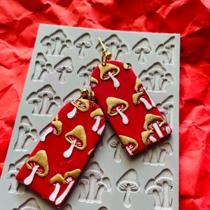May include: A pair of red polymer clay earrings shaped like a rectangle with a mushroom pattern in gold and white. The earrings are shown on a silicone mold with a mushroom pattern.