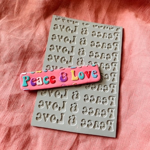 May include: A gray silicone mold with the words "Peace & Love" repeated multiple times. A pink and multi-colored clay piece with the words "Peace & Love" is sitting on top of the mold.