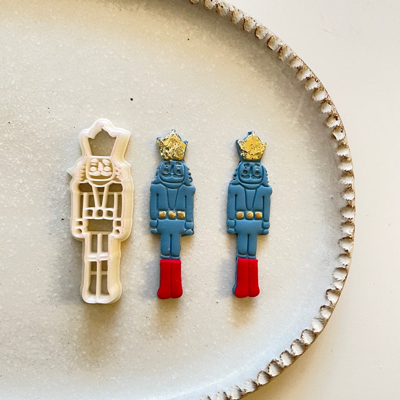 Pode incluir: Dois cortadores de biscoitos de quebra-nozes azul e dourado e um cortador de biscoitos de quebra-nozes de pl&aacute;stico branco em um prato branco.