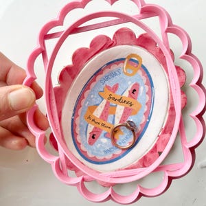 May include: A pink, scalloped-edge frame holds a sardine-themed illustration and a gold ring with a purple stone. The illustration features a sardine can design with the words "Sardines" and "In Pure Olive" on a blue and pink background.