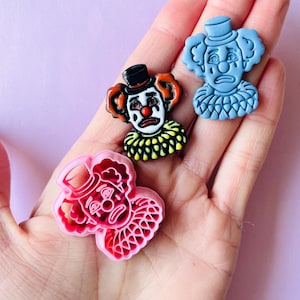 Pode incluir: Três pingentes de plástico com formato de rosto de palhaço em diferentes cores. Os pingentes são azuis, rosa e amarelos. Cada pingente tem uma expressão triste com um nariz vermelho e olhos pretos.