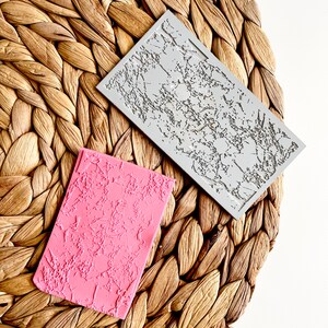 May include: Two silicone molds for creating textured surfaces. The mold on the left is pink and the mold on the right is gray. Both molds have a textured surface that resembles cracked concrete.