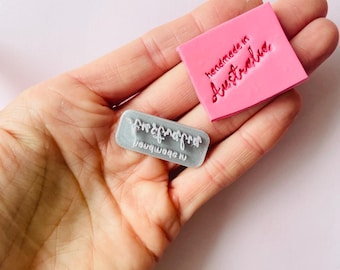 Op de afbeelding: Twee rubberen stempels, een roze en een grijze. De roze stempel zegt "handgemaakt in Australië". De grijze stempel zegt "gegraveerd door Nauquagon".