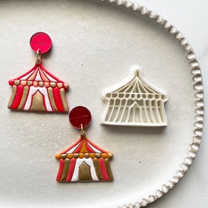 Könnte beinhalten: Zwei Paare rote, weiße und goldene Zirkuszelte Ohrringe. Die Ohrringe liegen auf einem weißen Teller mit einem gewellten Rand.