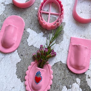 Op de afbeelding: Roze klei gereedschap, waaronder ovale en U-vormige uitsteekvormen, en een decoratieve ovale vaas met een hartontwerp en bloemen. De vaas is gevuld met kleine paarse en groene bloemen.