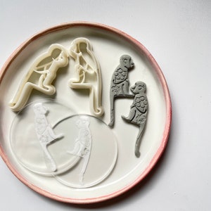 Pode incluir: Um conjunto de cinco cortadores de biscoitos em forma de macacos. Os cortadores são feitos de plástico branco e cinza. Os cortadores estão em um prato branco com uma borda rosa.