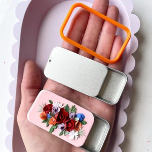 Könnte beinhalten: Zwei kleine Metalldosen mit Deckeln. Eine Dose ist mit einem rosa Hintergrund und einem bunten Blumendesign verziert. Die andere Dose ist schlicht silberfarben. Ein leuchtend orangefarbenes Plastikrechteck ist ebenfalls abgebildet.
