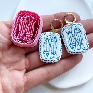 May include: A pink polymer clay fish-shaped mold and two pairs of earrings. The earrings feature a white base with blue fish designs and gold hoops. The mold has a pink, scalloped border and a fish design.