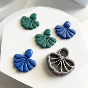 May include: Five clay earrings in the shape of a scallop shell. Two are green, two are blue, and one is gray. The gray earring is a mold for making the other earrings.