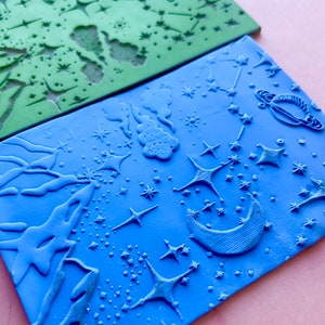 May include: Two silicone stamps with a celestial theme. The top stamp is green and features stars, a crescent moon, and a planet. The bottom stamp is blue and features stars, a crescent moon, a planet, and clouds.