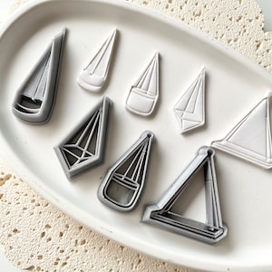 May include: A set of eight grey and white plastic cookie cutters shaped like sailboats. The cutters are arranged on a white plate with a lace pattern.