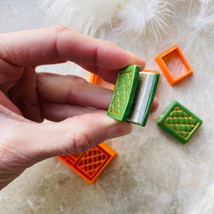 May include: A miniature green book with gold detailing, open to reveal blank pages. The book is held in a hand, and two orange boxes are visible in the background.