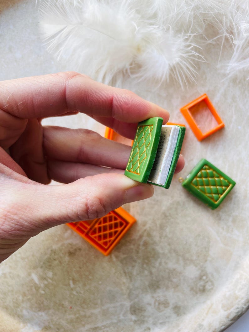 May include: A miniature green book with gold detailing, open to reveal blank pages. The book is held in a hand, and two orange boxes are visible in the background.