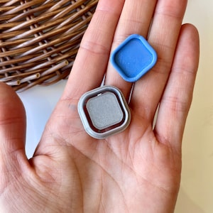 May include: Two small, square-shaped, decorative items. One is a light blue, plastic, square with a raised edge. The other is a silver, plastic, square with a raised edge and a textured center.