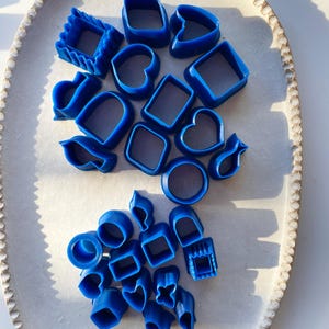 May include: An assortment of blue polymer clay cutters in diverse shapes, such as squares, hearts, and circles, are displayed on a white, oval ceramic tray. The cutters vary in size and design, ideal for crafting or baking activities.
