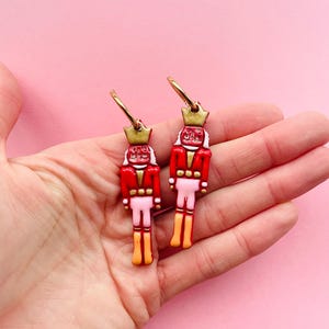 Pode incluir: Um par de brincos de argola dourados com figuras de quebra-nozes vermelhas, brancas e douradas penduradas neles. Os quebra-nozes usam jaquetas vermelhas, cal&ccedil;as brancas e chap&eacute;us dourados.