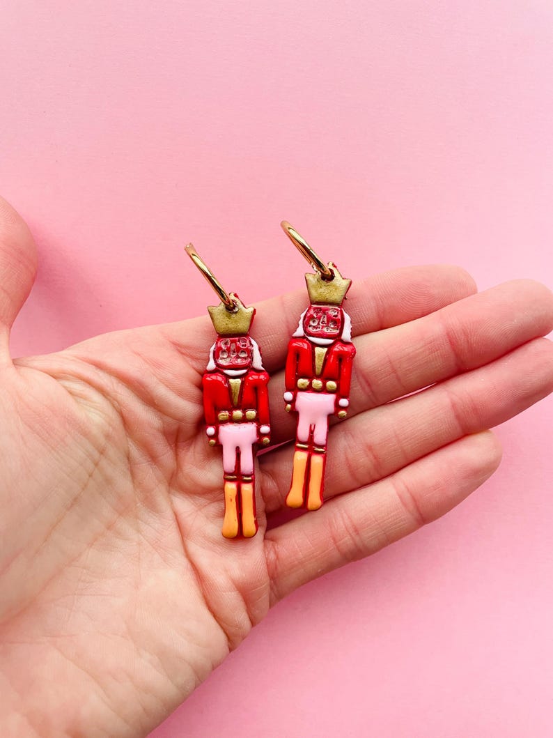Pode incluir: Um par de brincos de argola dourados com figuras de quebra-nozes vermelhas, brancas e douradas penduradas neles. Os quebra-nozes usam jaquetas vermelhas, cal&ccedil;as brancas e chap&eacute;us dourados.