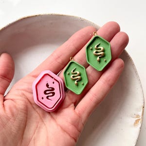 May include: Two hexagonal shaped charms with a gold snake design. One charm is pink and the other is green.