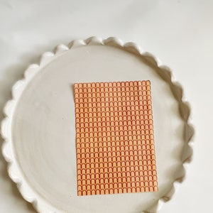 May include: A sheet of decorative paper with a repeating pattern of orange, yellow, and red arches. The paper is on a white ceramic plate with a scalloped edge.