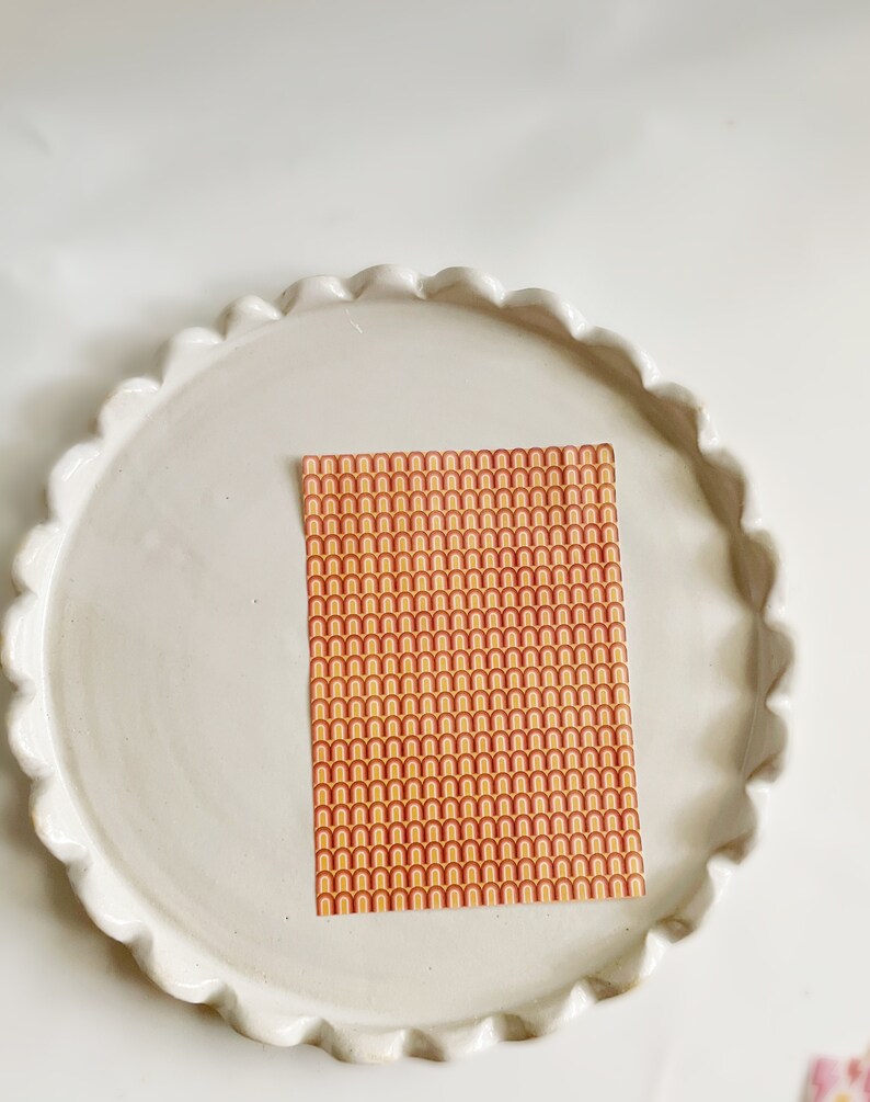 May include: A sheet of decorative paper with a repeating pattern of orange, yellow, and red arches. The paper is on a white ceramic plate with a scalloped edge.