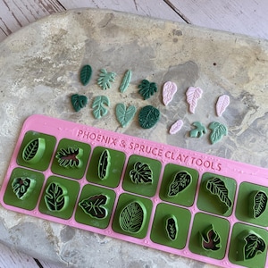 Puede incluir: Una colección de herramientas de arcilla con forma de hoja y hojas de arcilla terminadas en varios tonos de verde y rosa. Las herramientas están en una bandeja rectangular rosa con el texto "PHOENIX & SPRUCE CLAY TOOLS". Las hojas están dispuestas sobre una superficie gris claro.