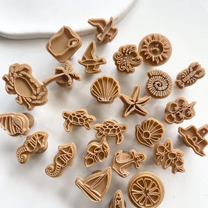 May include: A collection of tan-colored cookie cutters in various shapes, including a starfish, seashell, seahorse, turtle, and sailboat. The cutters are arranged on a white surface, perfect for creating beach-themed treats.