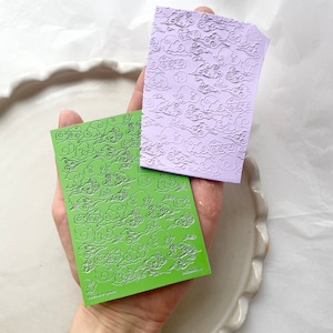 May include: Two sheets of green and purple silicone embossing mats with a repeating pattern of stylised clouds. The mats are designed for use with polymer clay, fondant, and other crafting materials.