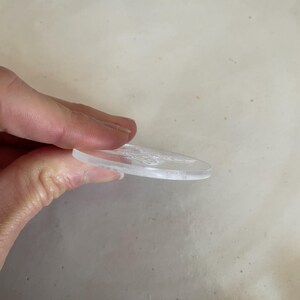 Puede incluir: Un disco redondo de acr&iacute;lico transparente con un dise&ntilde;o en relieve. El disco est&aacute; siendo sostenido por una mano.