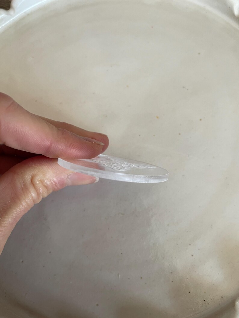 Puede incluir: Un disco redondo de acr&iacute;lico transparente con un dise&ntilde;o en relieve. El disco est&aacute; siendo sostenido por una mano.