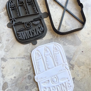 Puede incluir: Un juego de tres cortadores de galletas con forma de sardina. Los cortadores son negros y blancos, con la palabra "SARDINE" en una fuente curva. El diseño incluye tres peces dentro de una lata, una rodaja de limón y una pestaña.