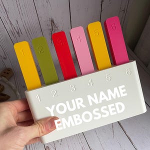 May include: A white rectangular holder with the text YOUR NAME EMBOSSED and numbers 1 through 6 printed on the front. Six colourful rectangular markers in yellow, olive green, red, light pink, yellow, and dark pink are inserted, each labelled 1-6.